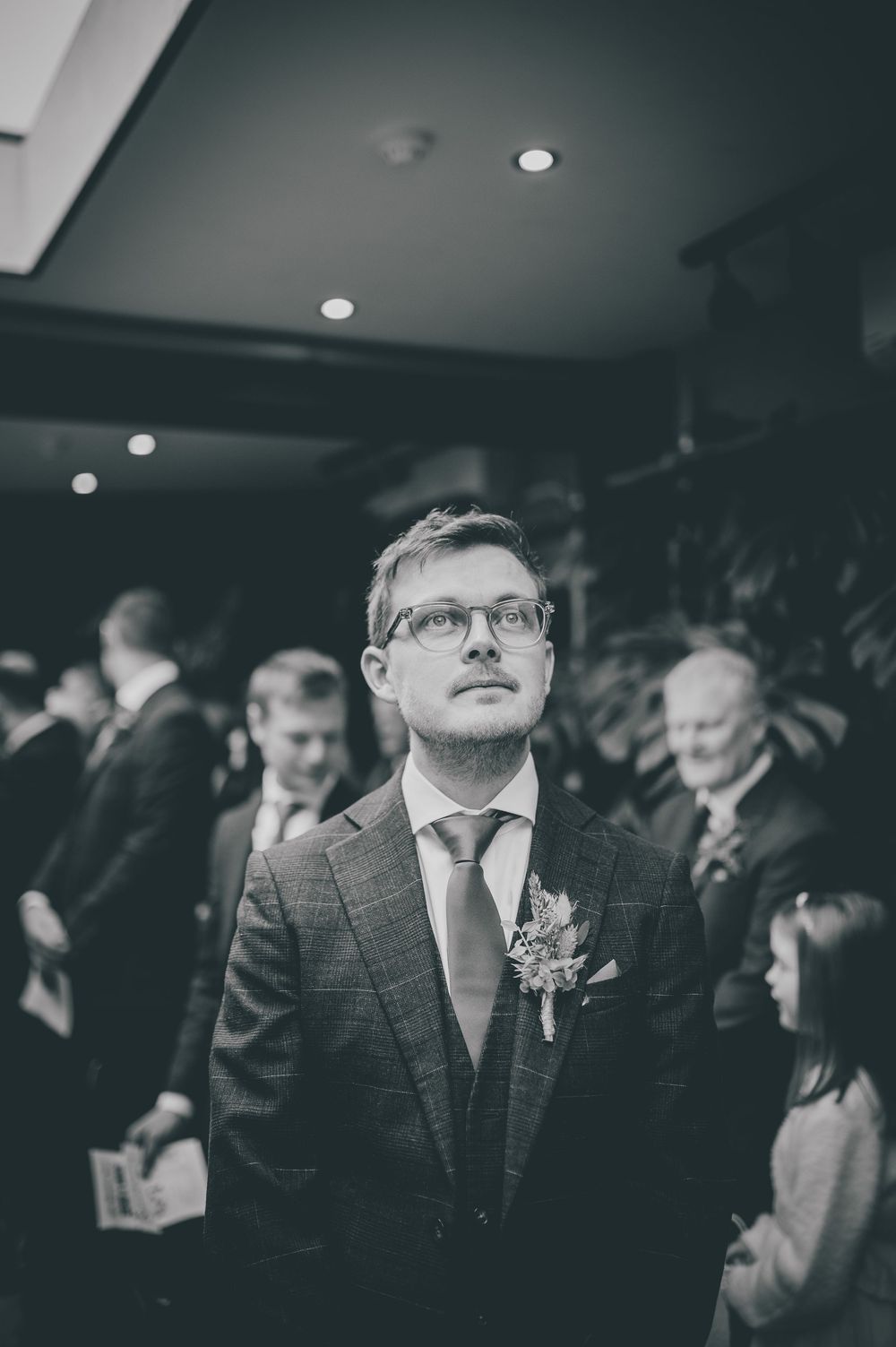 Black and white photo of a well-dressed guest wearing glasses and a boutonniere at a formal indoor event.