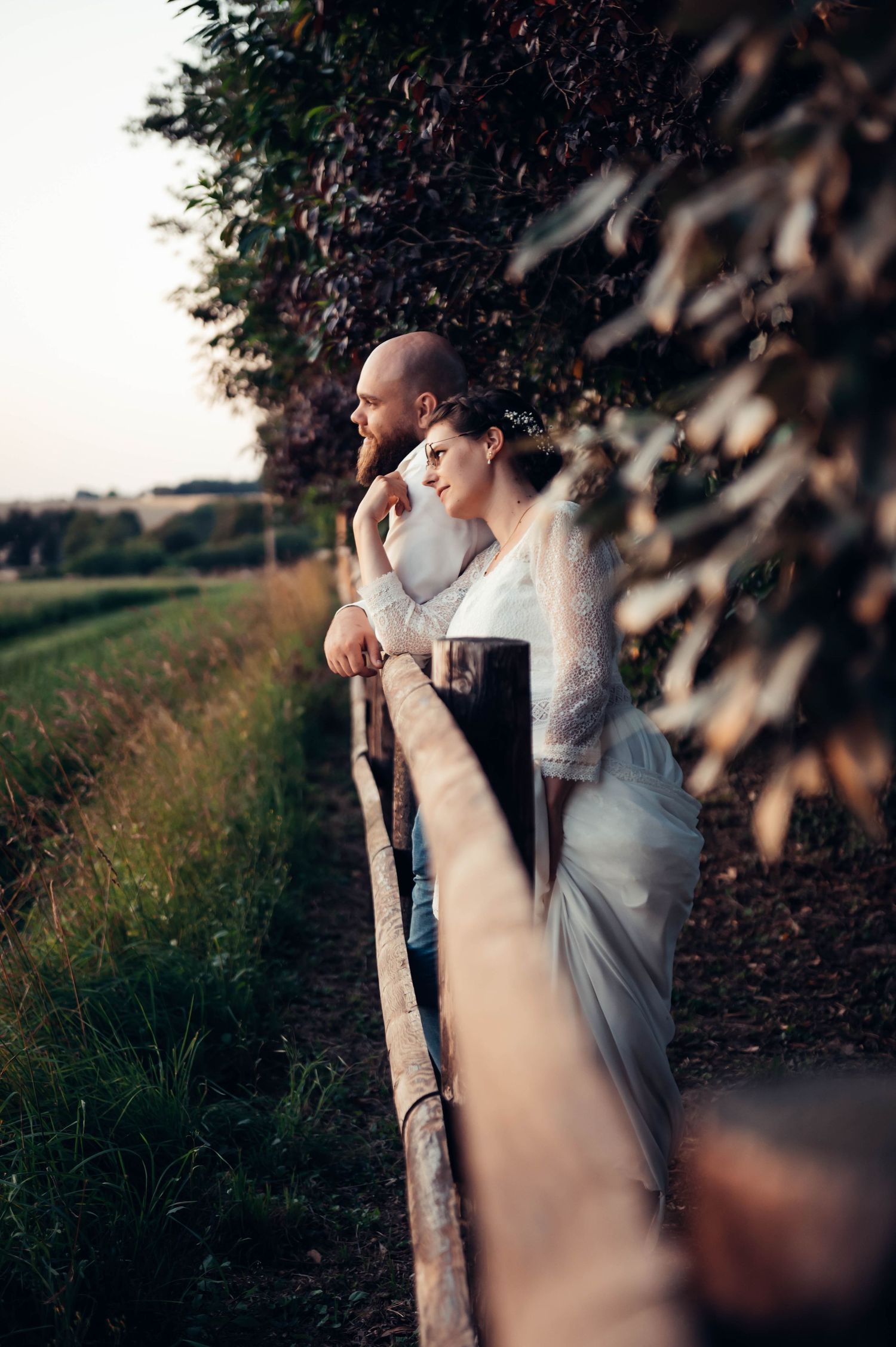 Couple de mariés le long d’une barrière en bois au coucher du soleil, séance photo romantique en campagne bretonne.