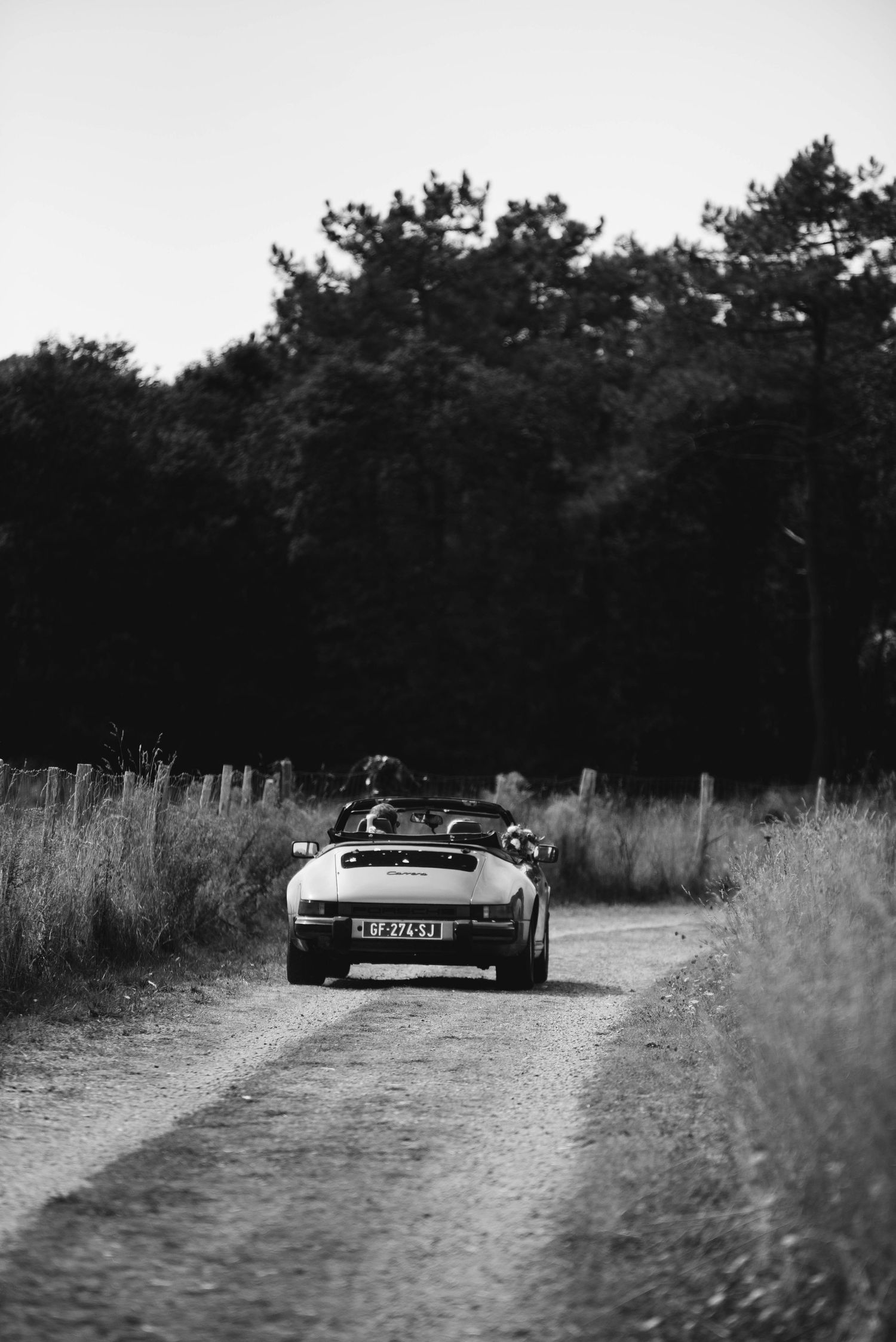 Voiture vintage roulant sur un chemin de campagne lors d’un mariage, photo en noir et blanc.