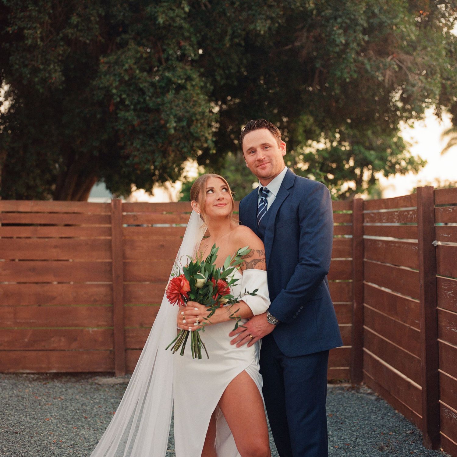 Bride and groom stand together in traditional wedding attire. The bride is holding a bouquet of autumn florals and looking at the groom.