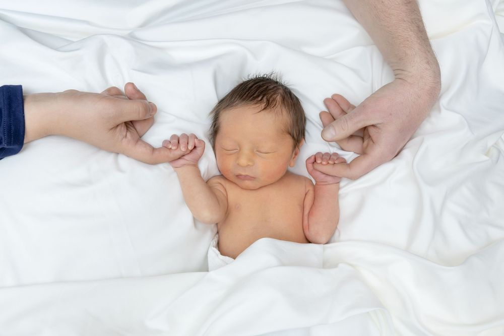 A sleeping newborn baby lies peacefully on white bedding while being tenderly touched by caring hands.