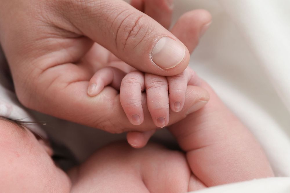 A close-up photo of a tiny baby hand gently grasping an adult finger against a soft white background.