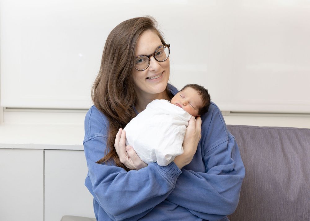 A person in a blue sweater gently cradles and gazes at a sleeping newborn baby wrapped in white.
