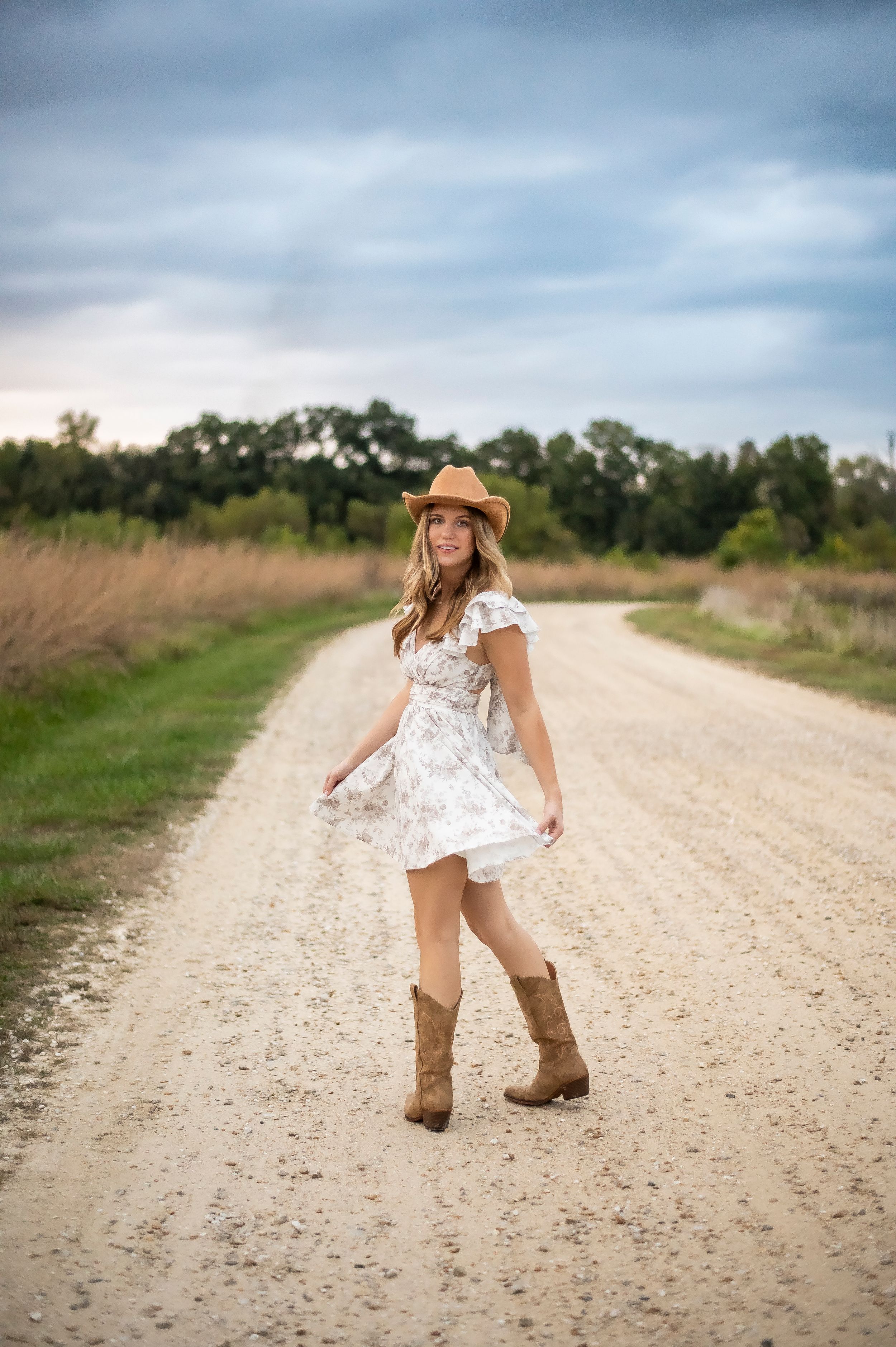 Young woman in white floral dress, cowboy hat, and boots twirling on a rustic dirt road.