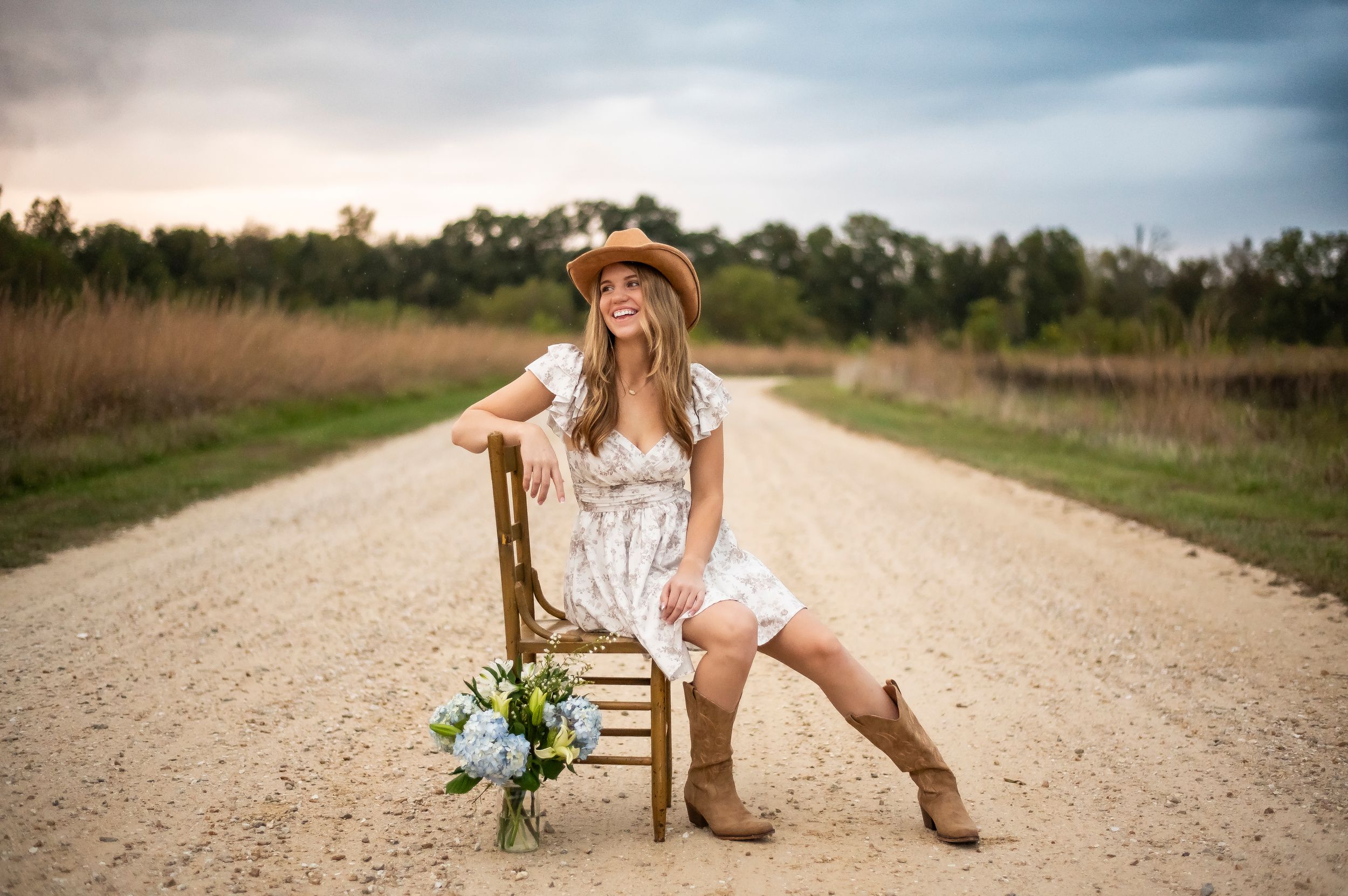 Woman in white dress and cowboy boots sits on a chair on a dirt road with wildflowers.