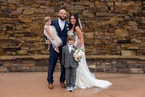 Bride and groom posing with two young children in front of a rustic stone wall at wedding.