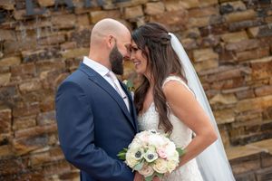 Bride and groom gazing lovingly at each other holding a white floral bouquet against a stone wall.