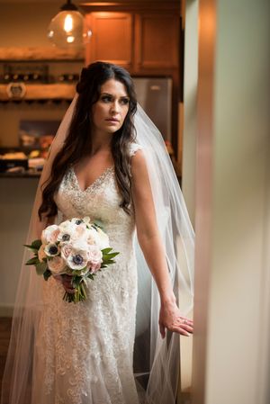 Bride in lace gown and veil holds white and blush bouquet in indoor portrait.