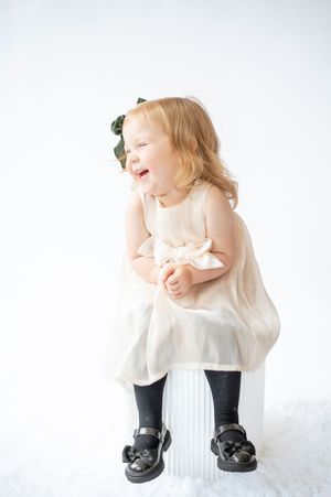 Cute toddler girl in cream tulle dress with green bow laughing on white cube.