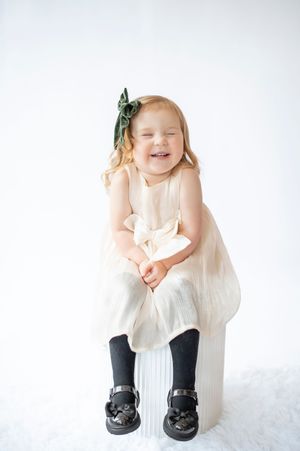 Happy little girl in cream dress with green hair bow laughing in white studio.