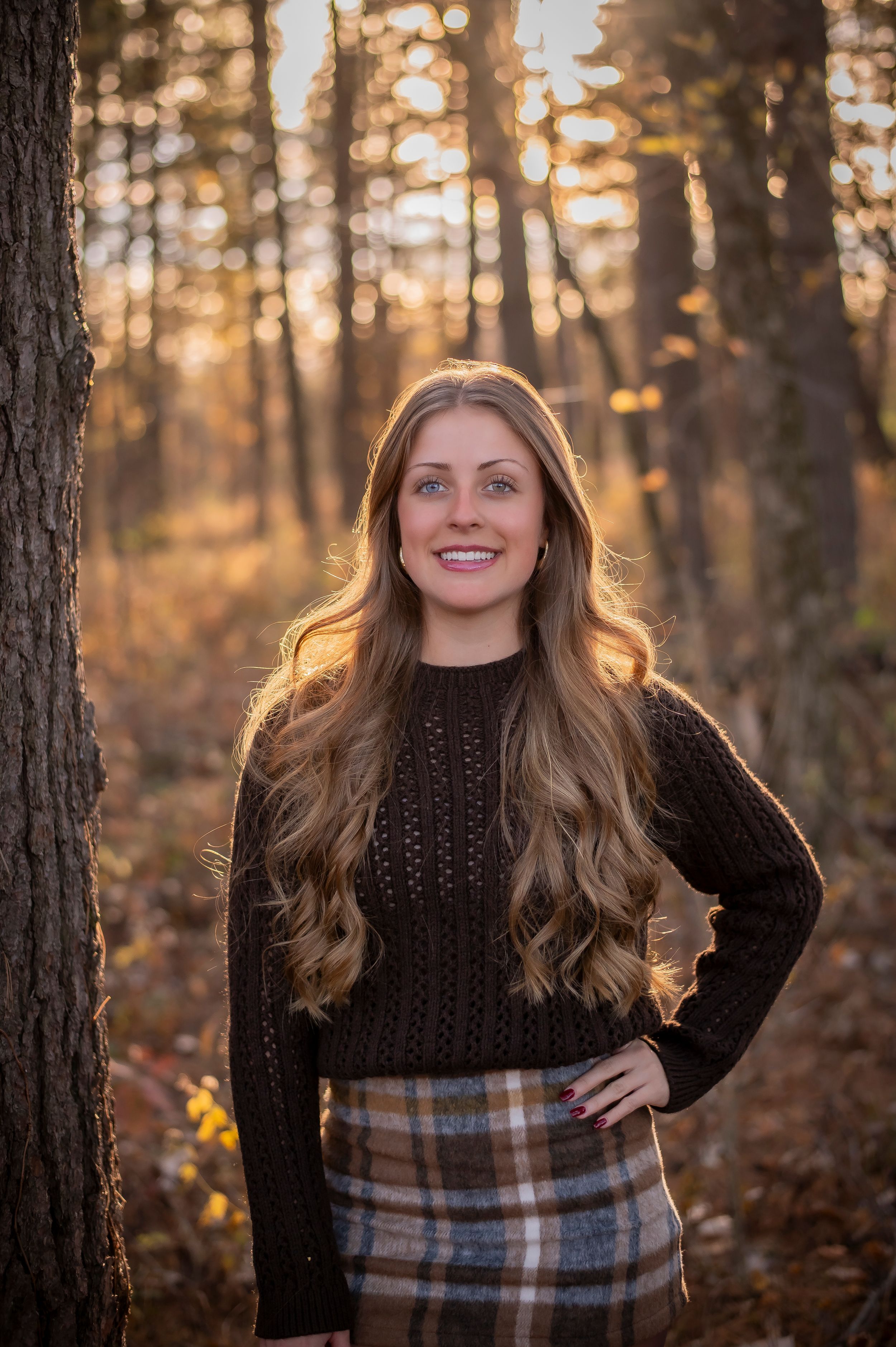 Young woman with long wavy hair smiling in autumn forest wearing cozy sweater for senior portrait.