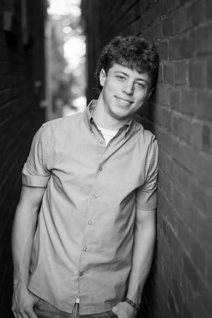 Smiling teenage boy leaning against brick wall in black and white portrait photo.