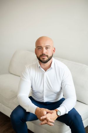 Bald man with beard in white shirt sitting on white couch with hands clasped.
