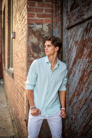 Stylish young man in light blue shirt and white pants posing by rustic brick wall.