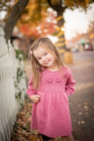 Smiling toddler girl in a pink knit rose dress posing outdoors in autumn leaves by a fence.