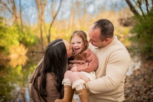 Parents kissing their laughing toddler daughter outdoors near a colorful autumn creek.