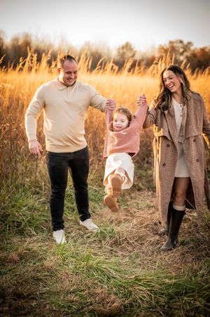 Happy family swinging their toddler daughter between them in a sunlit autumn field.