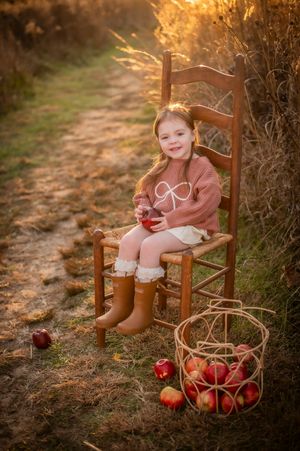 Adorable little girl in pink sweater sits on wooden chair with apples at sunset.