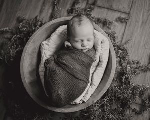 Peaceful newborn baby swaddled in wrap sleeping in wooden bowl, black and white photo.