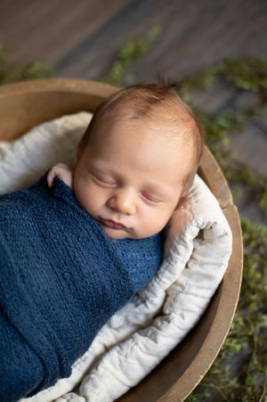 Sleeping newborn baby swaddled in a navy blue wrap resting in a wooden bowl with white linen.