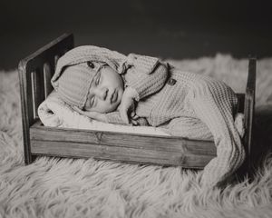 Black and white photo of a sleeping newborn in a knit outfit on a tiny wooden bed.