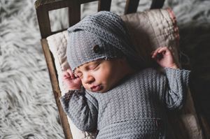 Newborn baby sleeping in a gray knit romper and hat on a rustic wooden bed.