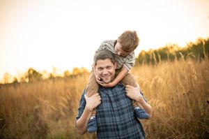 Man carries smiling boy on shoulders in a golden wheat field at sunset.