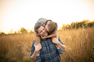 Young boy hugs man from behind in a warm golden field at sunset.