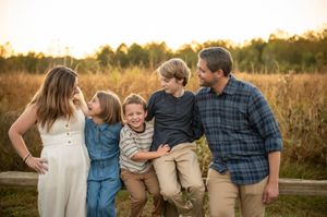 Family of five laughs together leaning on a fence in a sunlit autumn field.