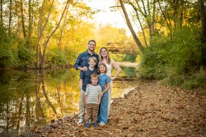 Family of five poses by a reflective creek surrounded by colorful autumn trees.