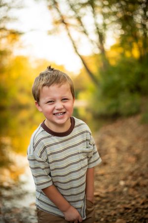 Cheerful blonde boy smiles broadly on a leaf-covered path by a sunlit creek.