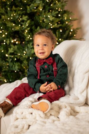 Adorable baby boy in green holiday outfit sitting near lit Christmas tree.