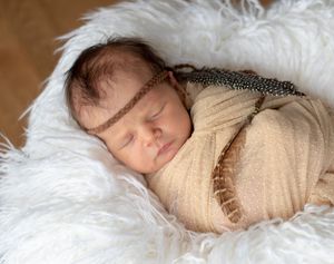 Sleeping newborn baby swaddled in cream wrap with feather headband resting in fluffy white basket.