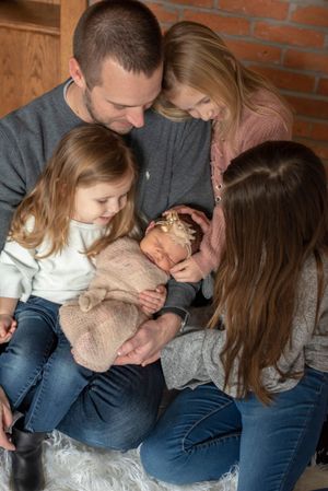 Family of five gathered closely together admiring their swaddled newborn baby indoors.