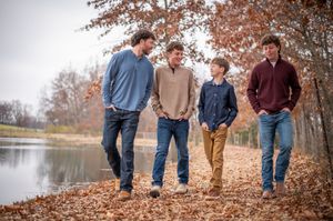 Four males walking together laughing among fallen leaves near tranquil lakeside setting.