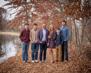 Family of six posing together among fall foliage near pond in wooded outdoor setting.