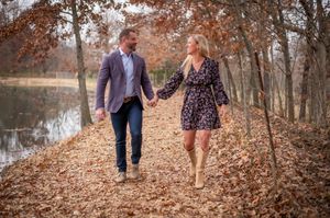 Couple walking hand in hand through autumn leaf-covered path beside serene pond.