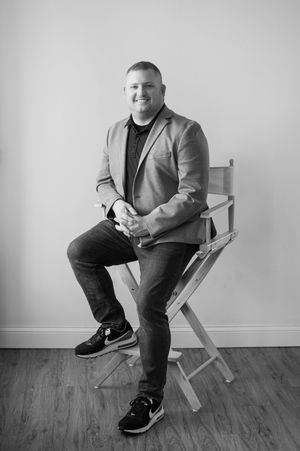 Man in blazer sitting on director's chair smiling in black and white professional portrait photo.