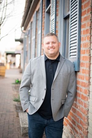 Smiling man in gray blazer leaning against brick wall outdoors in professional headshot.