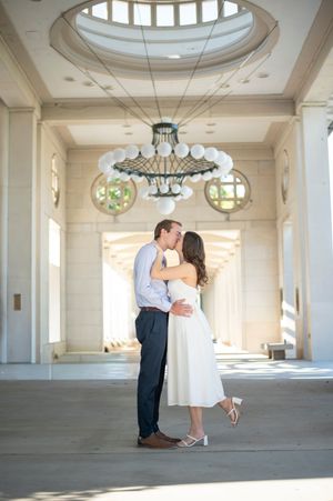 Couple sharing romantic kiss under elegant chandelier in grand architectural hall.