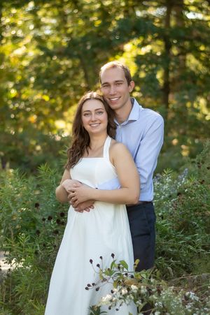 Smiling couple embraces in a lush green garden, woman in white dress, man in blue shirt.