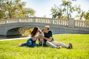 Couple sits on grass with their dog near a stone bridge in a sunny park.