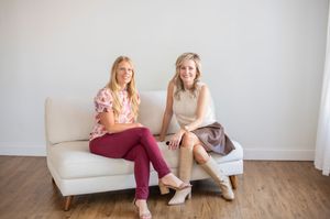 Two smiling women seated together on a white sofa in a bright minimalist studio.