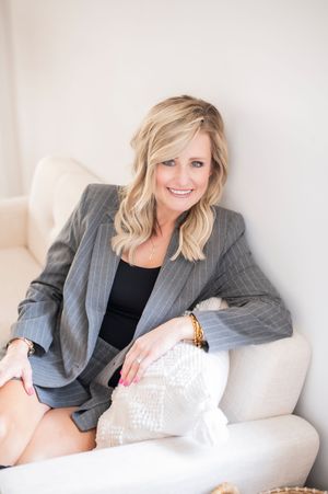 Confident blonde woman in gray pinstripe blazer sitting on white sofa smiling.