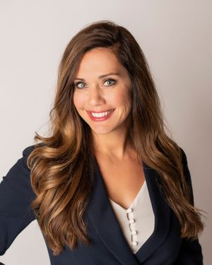 Professional headshot of a brunette woman in a navy blazer smiling confidently against a neutral background.