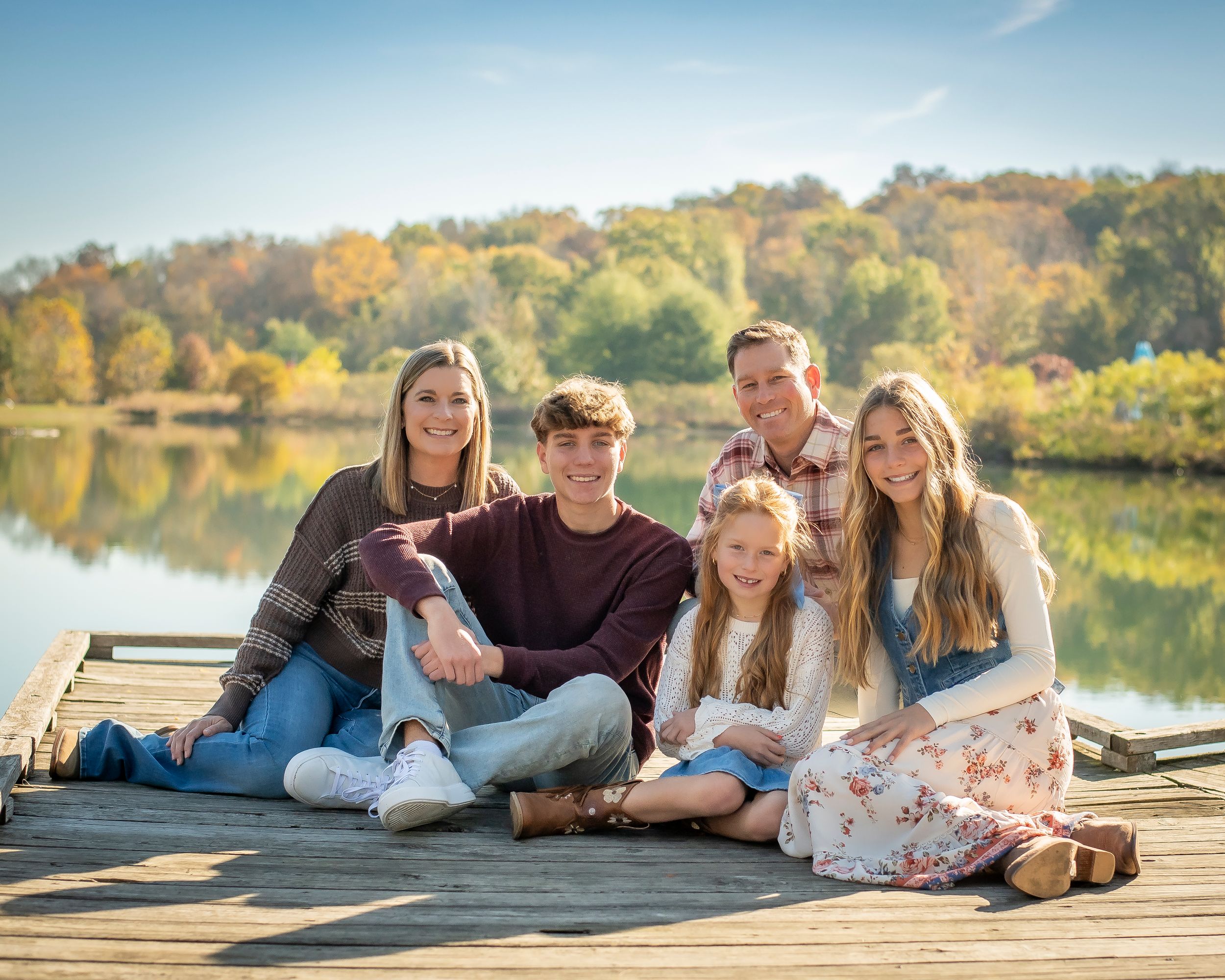 Family of six posing on wooden dock by scenic lake during colorful autumn outdoor portrait session.