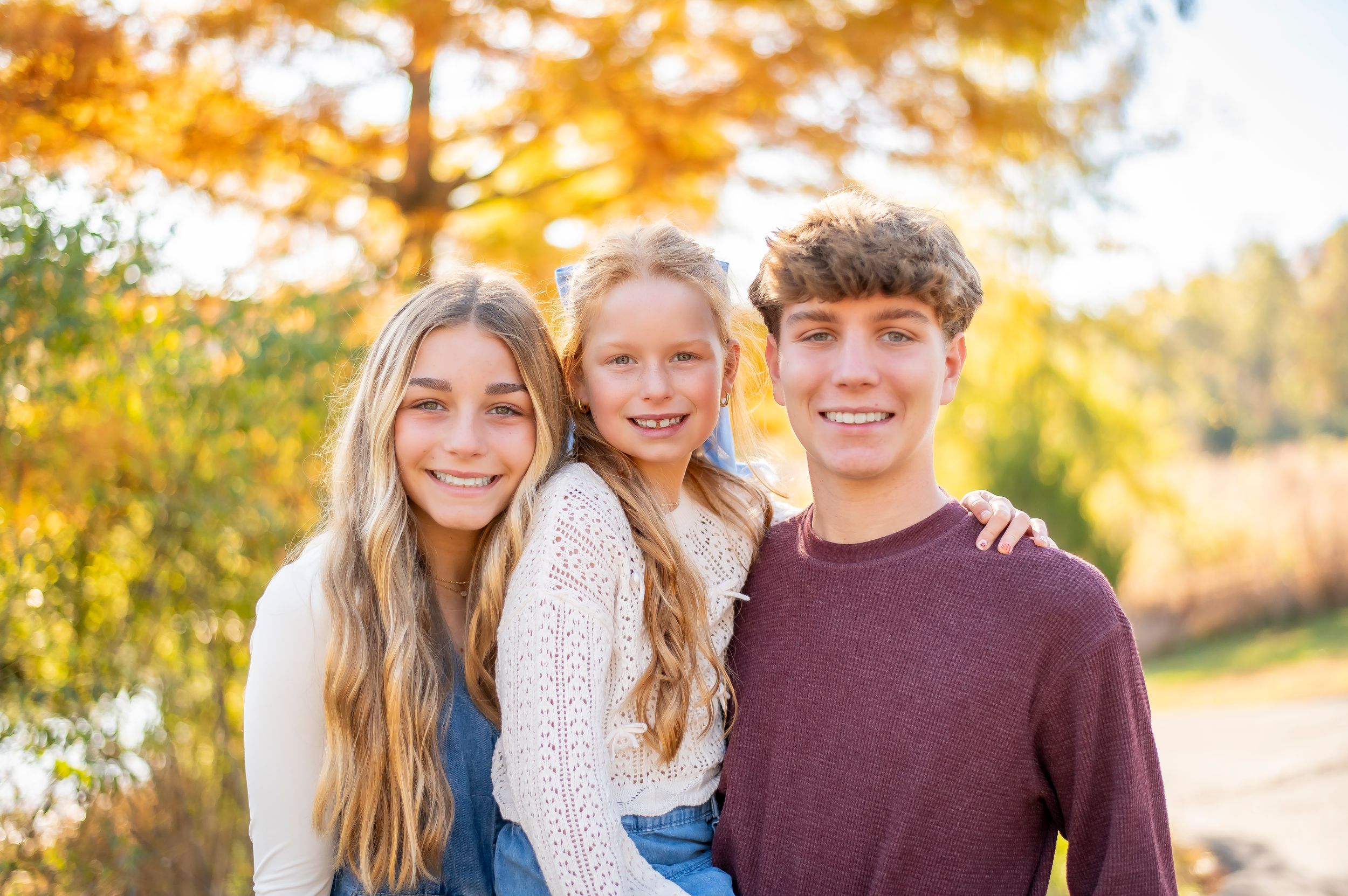 Three siblings smiling together outdoors with vibrant autumn foliage in warm fall family portrait.