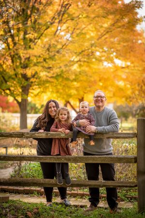 Happy family of four poses by wooden fence among vibrant fall foliage trees.