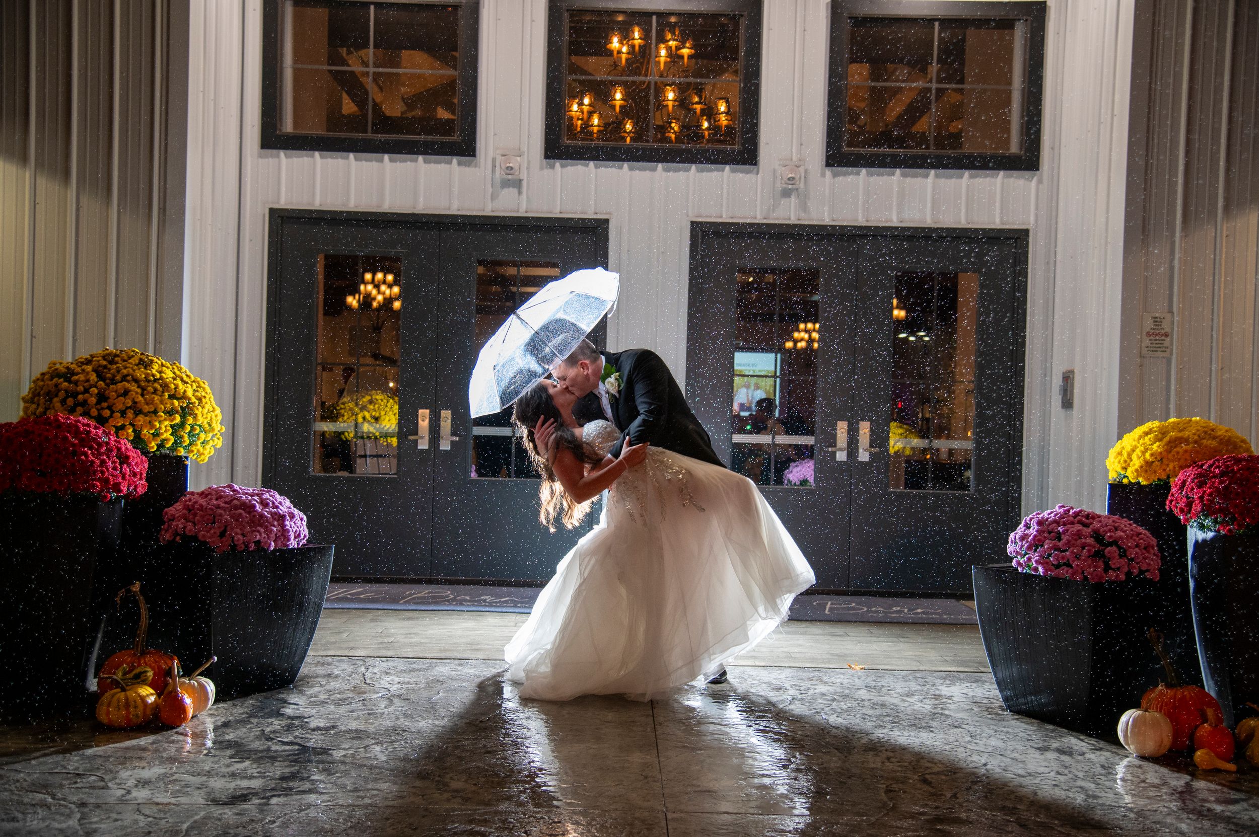 Bride and groom sharing romantic dip kiss under umbrella outside elegant venue on rainy wedding night.
