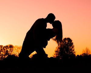 Romantic silhouette of couple kissing at sunset with warm orange sky background.